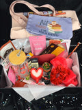 Gift box with various items including a tote bag, snacks, and a candle on a dark background.